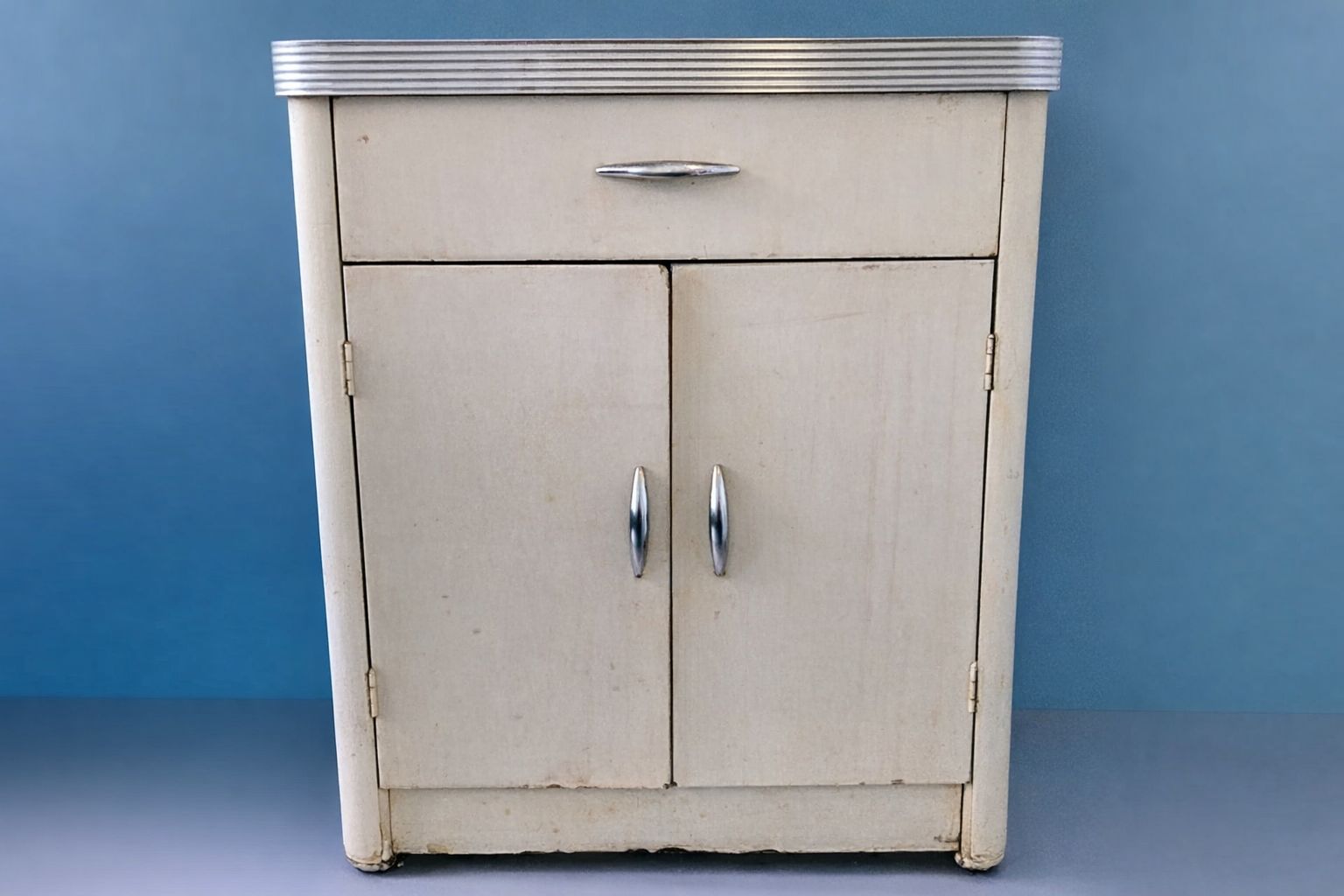 Wit vintage keukenkastje met zilverkleurige handgrepen tegen een blauwe wand.